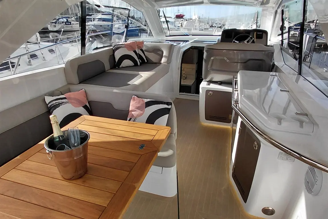 Luxury boat interior featuring a wooden table, champagne in a bucket, plush seating, and views of docked yachts.